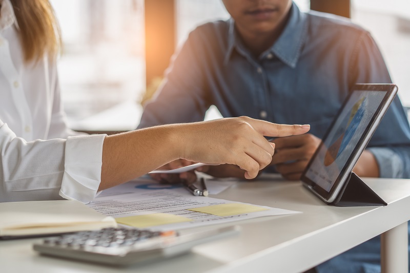 At a table, two people were sitting. A woman's hand points to the tab placed on the table where the business report is displayed.